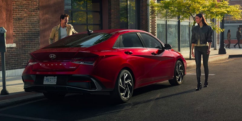 Un Hyundai Elantra rojo estacionado en una calle de la ciudad, con un hombre mirando dentro del maletero y una mujer caminando a su lado.