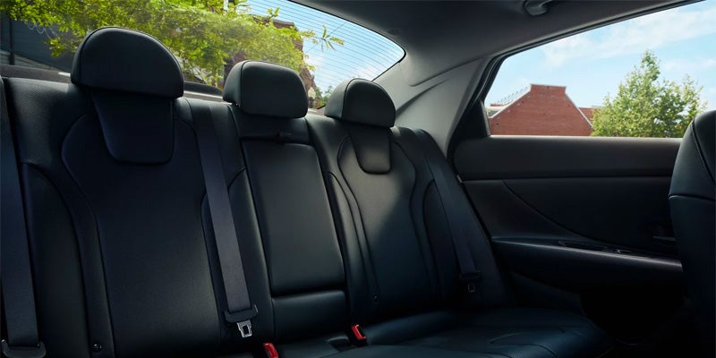 Vista interior de un coche con tres asientos traseros de cuero negro y una vista soleada de árboles y edificios de ladrillo a través de la ventana trasera.