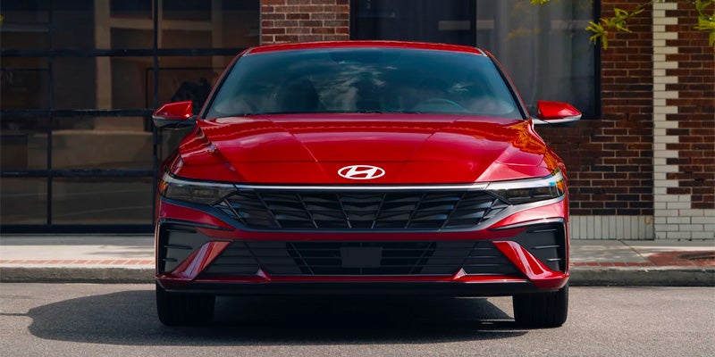 Vista frontal de un Hyundai Elantra rojo brillante estacionado en una calle frente a un edificio de ladrillo.