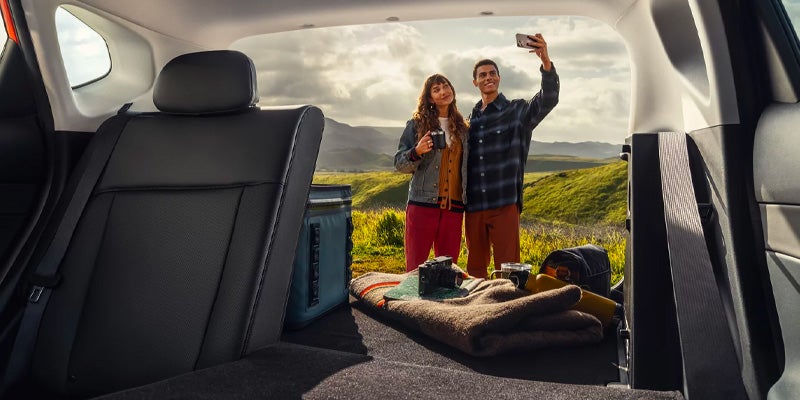 Vista desde la apertura del maletero de un coche de una pareja haciéndose un selfie con un pintoresco paisaje verde y montañoso de fondo.