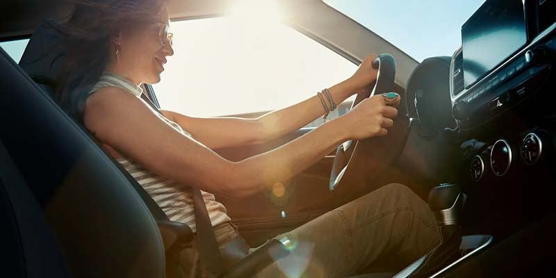 Una mujer con gafas de sol y una camisa de rayas conduce un coche, sonriendo, con la luz del sol entrando a raudales por el parabrisas.
