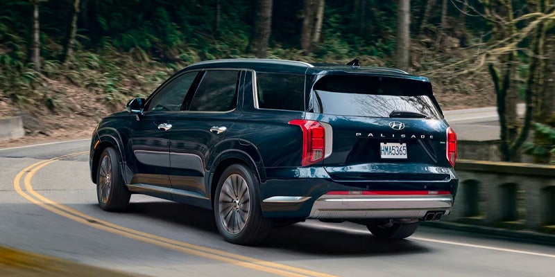 Vista trasera de un SUV Hyundai Palisade azul oscuro tomando una curva en una carretera bordeada de árboles.