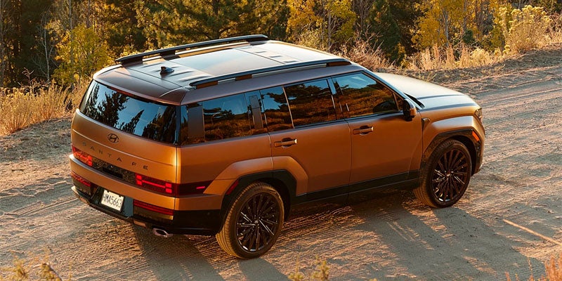 Vista trasera de un nuevo SUV Hyundai Santa Fe de color dorado terroso con ruedas negras circulando por un camino de tierra.