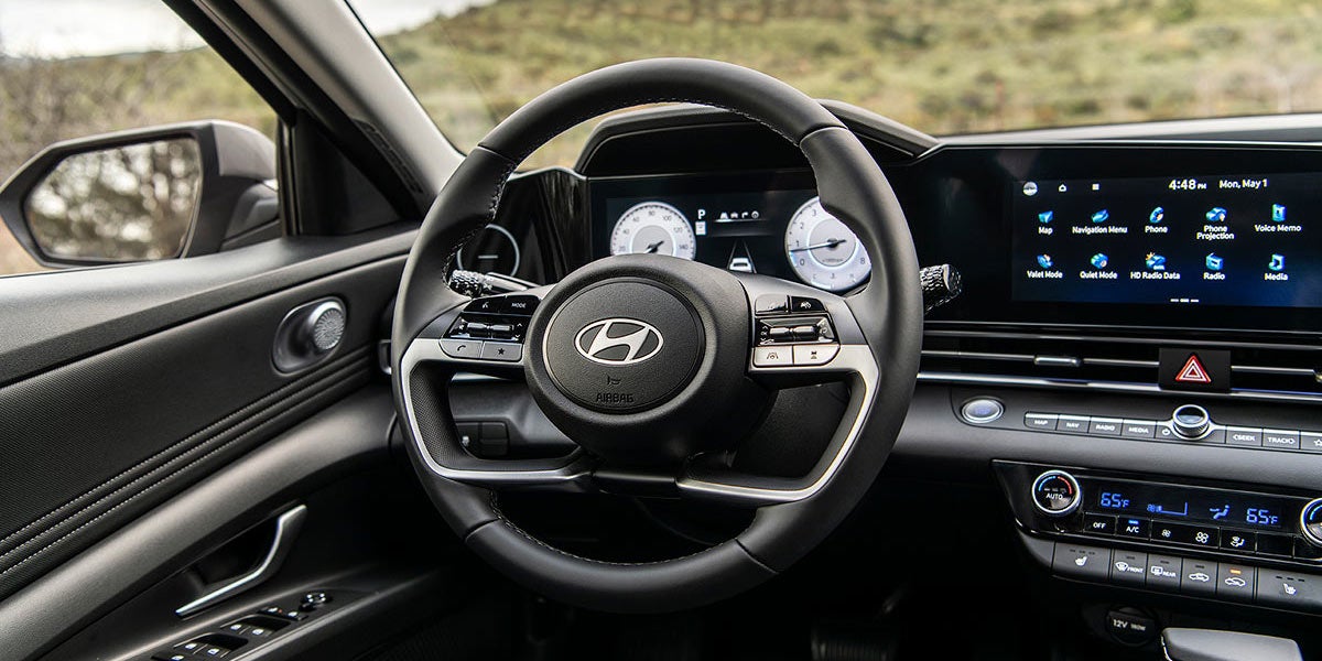 Interior shot of a dark-colored car's dashboard with a steering wheel showing the Hyundai logo, screens, and controls.