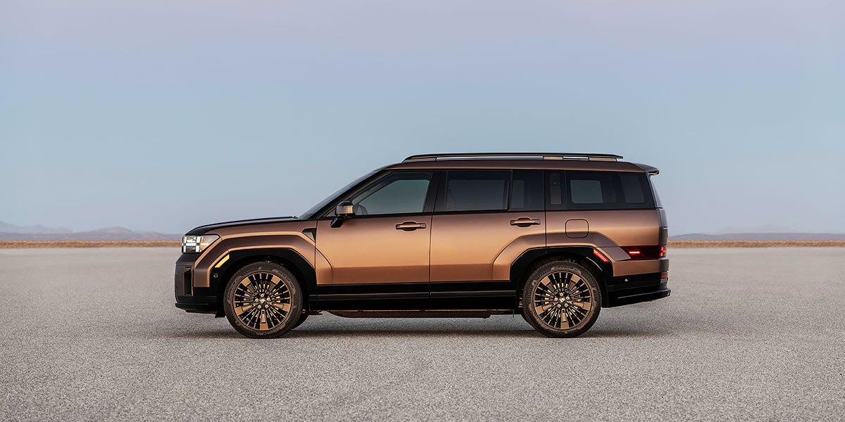 Un SUV de color bronce con parrilla rectangular y faros en forma de H está estacionado en una carretera con montañas al fondo.